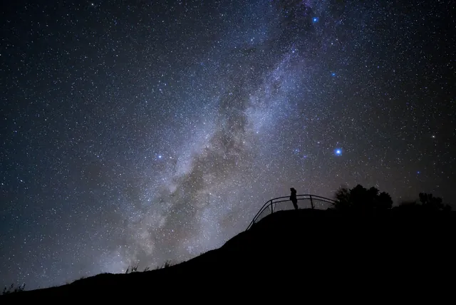 A silhouetted figure stands at a fenced overlook with the milky way arching above