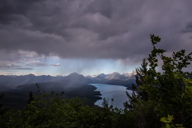 Trees in the foreground, mountains clouds, and a lake in the background.