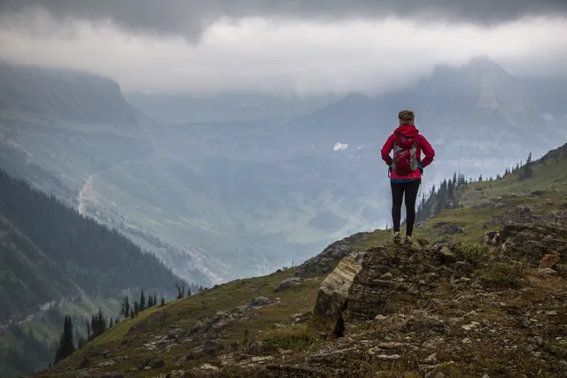 A hiker in red takes in a sweeping landscape cloaked in grey clouds.